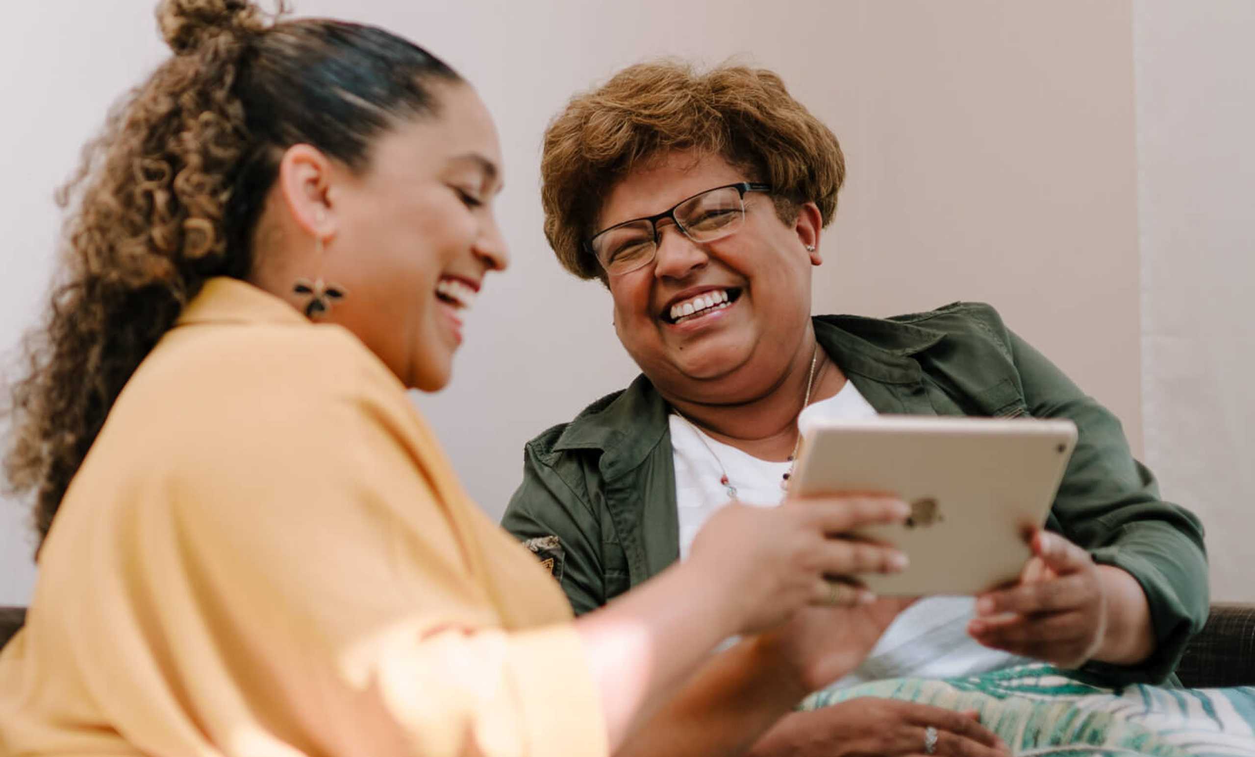 Two women sit close together holding an iPad - one hand each on the tablet. They are smiling and beaming, like old friends catching up.
