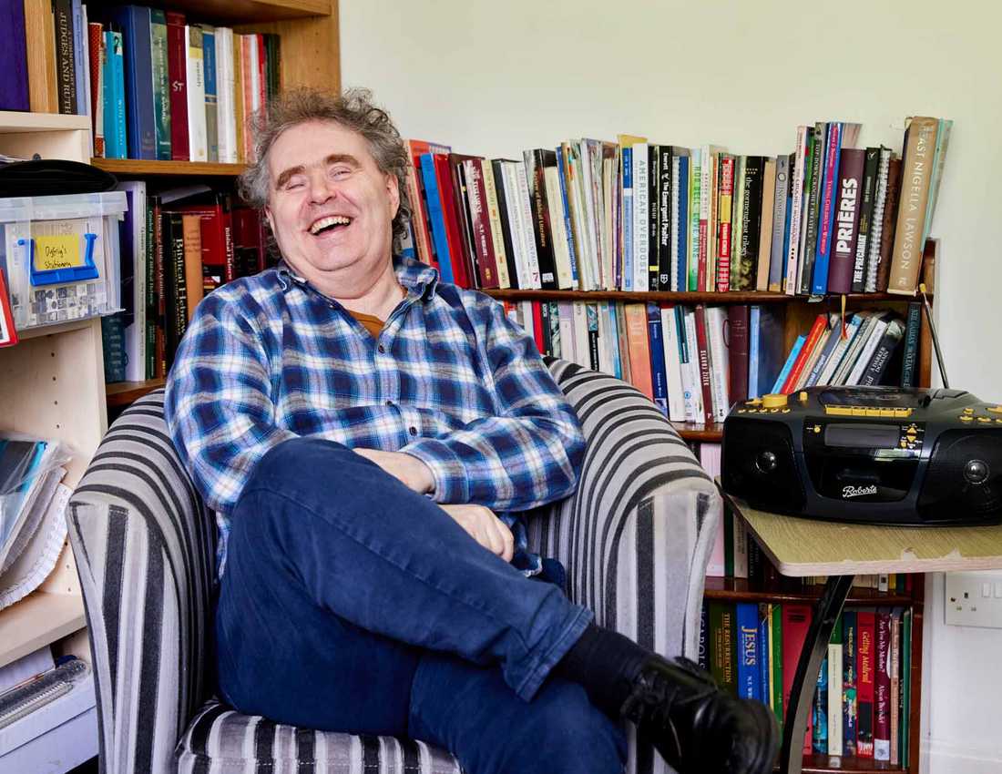 BWBF service user Dyfrig sits in a chair in a room with bookshelves. He is laughing and his Concerto 3 radio from BWBF is on a table next to him
