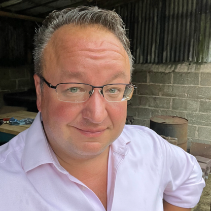 Alex Froom, Trustee for BWBF, smiling at the camera while standing in a workshop setting. He is wearing glasses and a pale pink shirt, with short greying hair, and a brick wall and wooden beams visible in the background.