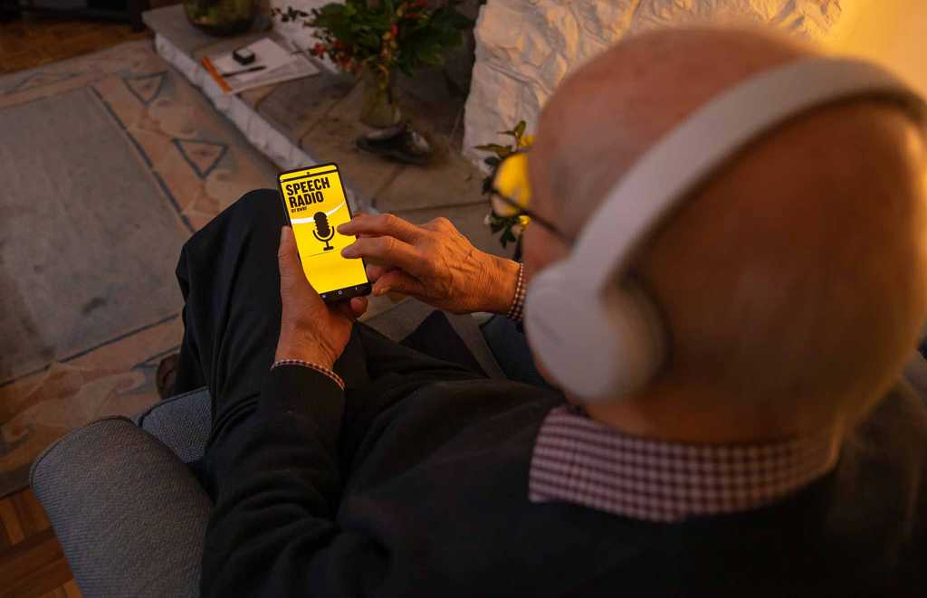 A man is browsing BWBF's Speech Radio app in a dimly lit room. He is wearing headphones.