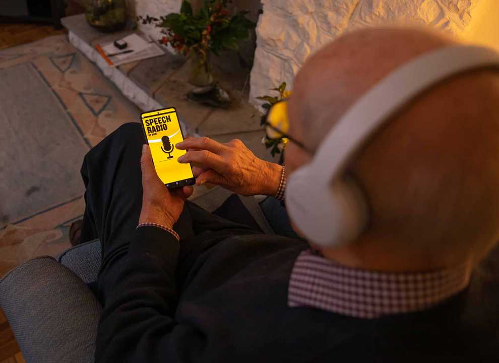A man is browsing BWBF's Speech Radio app in a dimly lit room. He is wearing headphones.