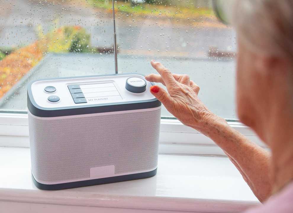 The Relish Radio sits on a windowsill as a user operates it. The radio is grey and white, with on/off button, four preset buttons and a volume control on top.