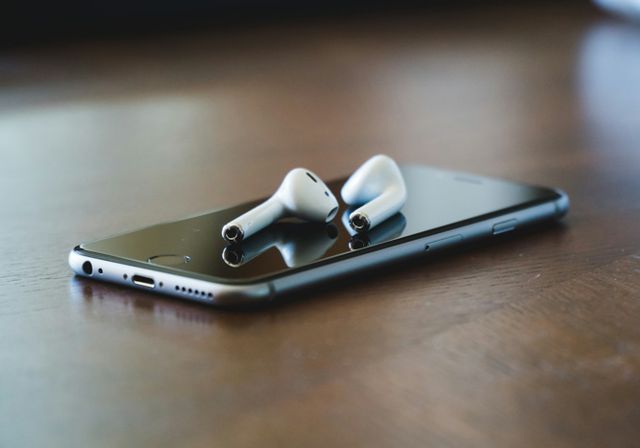 A mobile phone lies on black table surface. On top is a pair of cordless in-ear headphones.