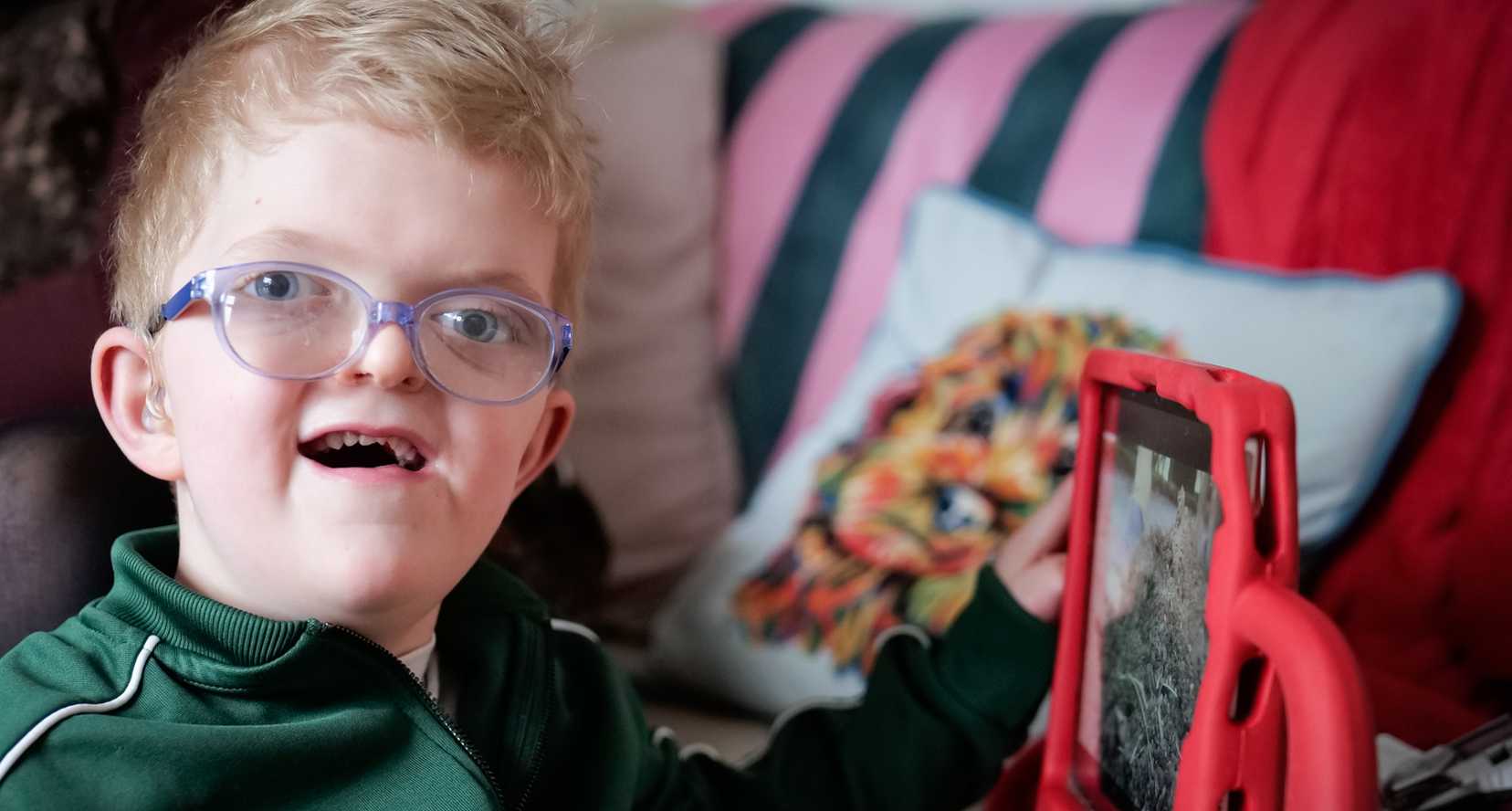 Chip, a young blonde boy, smiles broadly to camera as he explores his iPad from Visably.