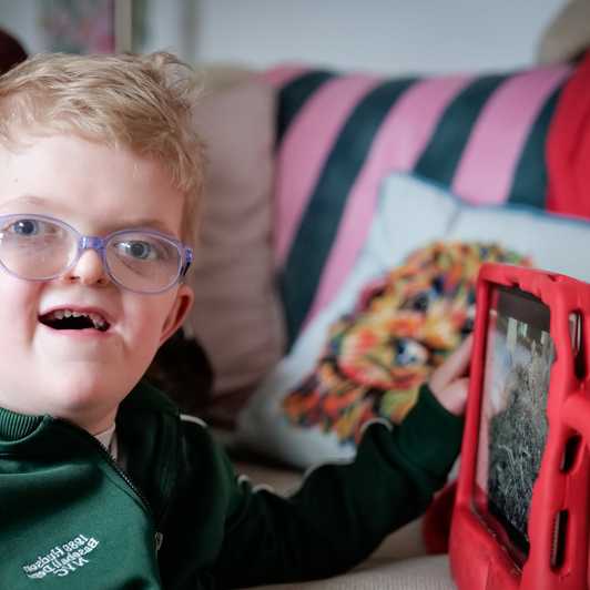 Chip, a young blonde boy, smiles broadly to camera as he explores his iPad from Visably.