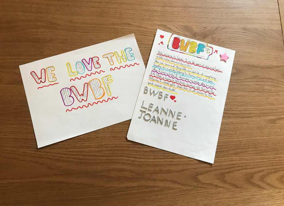 A colourful handwritten sign and letter lie expressing support for BWBF side by side on a wooden surface. The sign reads 'We love BWBF' and the letter sits next to it, signed off with a BWBF and a heart.
