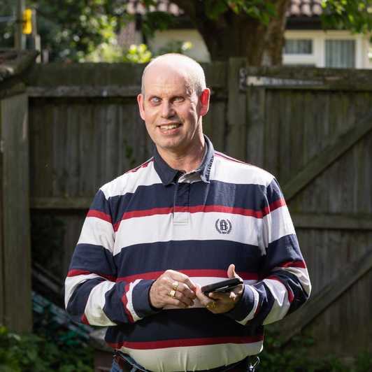 Brian, one of BWBF's app users, stands smiling in his garden as he holds his mobile phone.