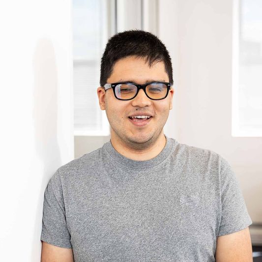 Suresh Kuwar, Web Developer for BWBF, is standing in an office space and smiling. He is wearing a grey t-shirt and glasses and has short, dark hair.