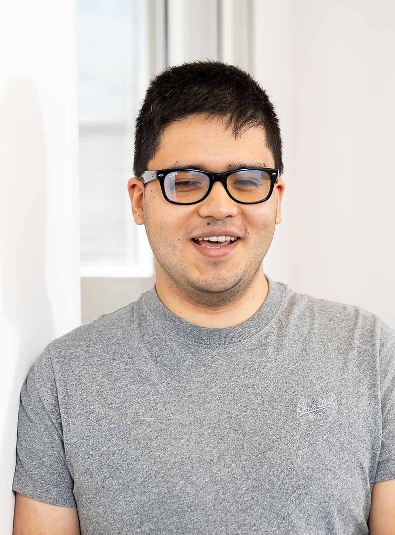 Suresh Kuwar, Web Developer for BWBF, is standing in an office space and smiling. He is wearing a grey t-shirt and glasses and has short, dark hair.