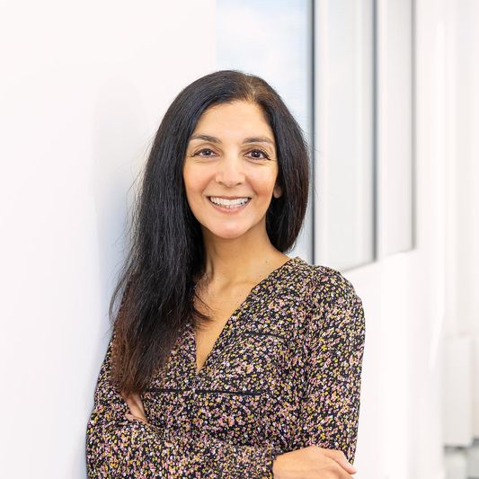 BWBF Communcations & Data Assistant Divya Kohli pictured in an office. She has her arms crossed and is smiling and wearing a brown floral dress.