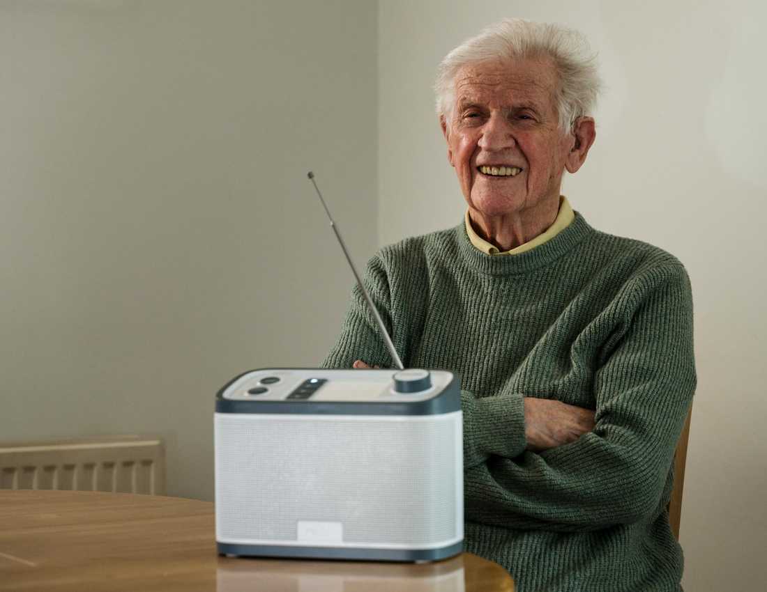 Thomas sits at a table smiling with his Relish radio from BWBF in front of him. His arms are folded.