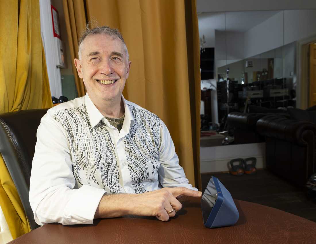 Paul Quigley, musician and performer. Paul lives with sight loss and is a beneficiary of BWBF. He is seated, smiling, as he engages with an Echo Show device on the table in front of him.