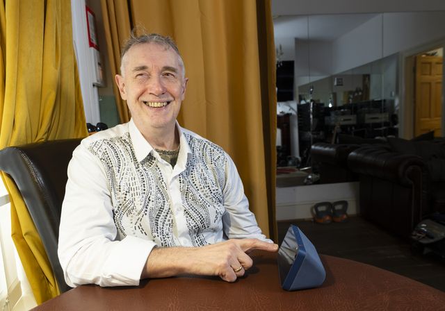 Paul Quigley, musician and performer. Paul lives with sight loss and is a beneficiary of BWBF. He is seated, smiling, as he engages with an Echo Show device on the table in front of him.
