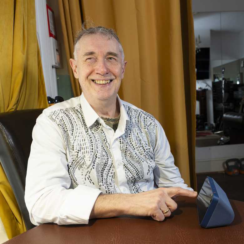 Paul Quigley, musician and performer. Paul lives with sight loss and is a beneficiary of BWBF. He is seated, smiling, as he engages with an Echo Show device on the table in front of him.