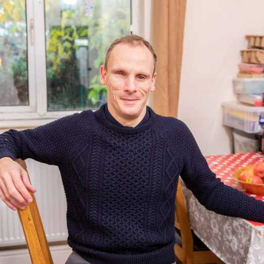 Jason, a user of BWBF's apps, sits at a table with a red checked table cloth. He is smiling and wearing a dark jumper.