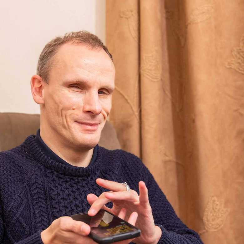 Jason is sitting on an armchair with a mobile phone in his hand. He is smiling as he navigates BWBF's app.