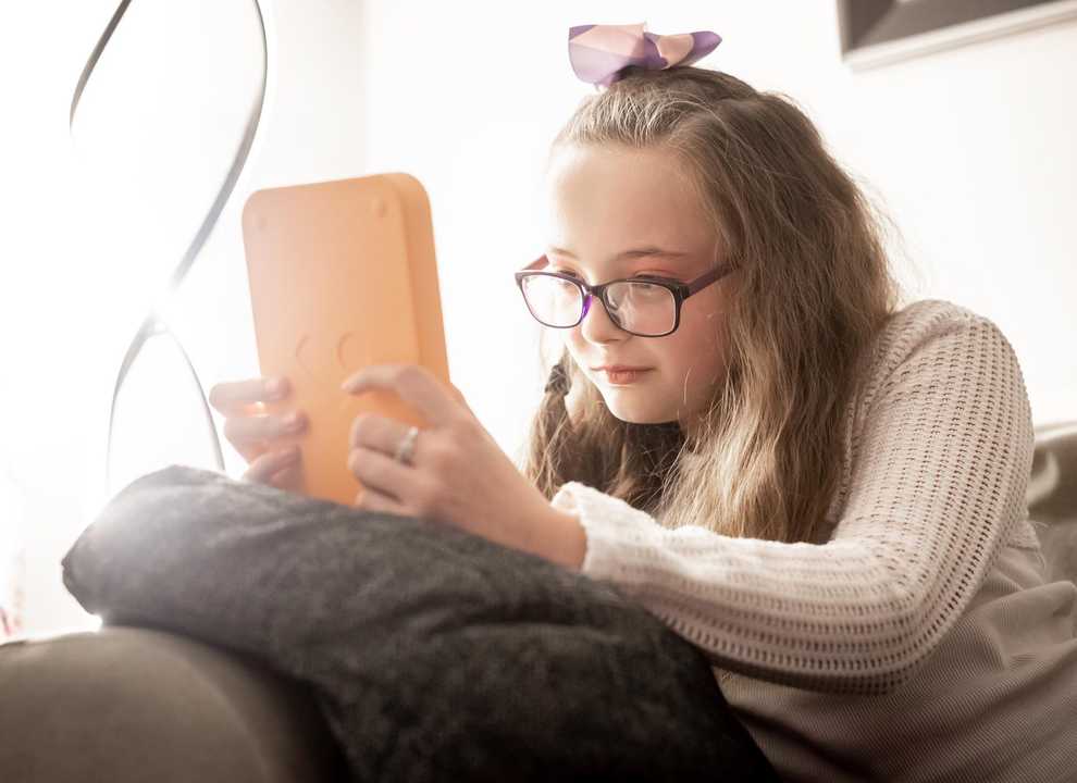 Annabel sits on her sofa reading her Voxblock audio book player. She wears glasses and has a bow in her hair.