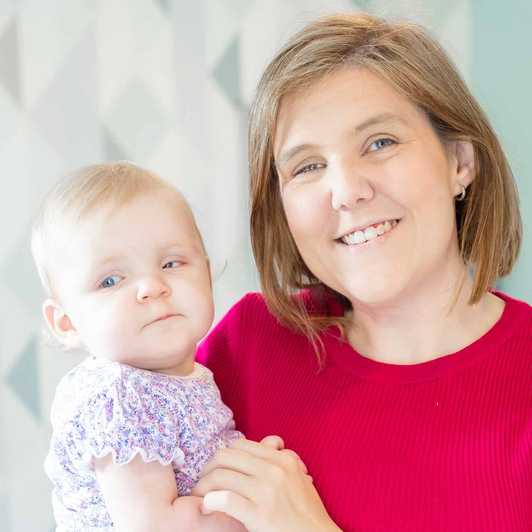 Young mother Laura smiles as she holds her baby daughter Daphne in her arms. She is wearing a vibrant red top.