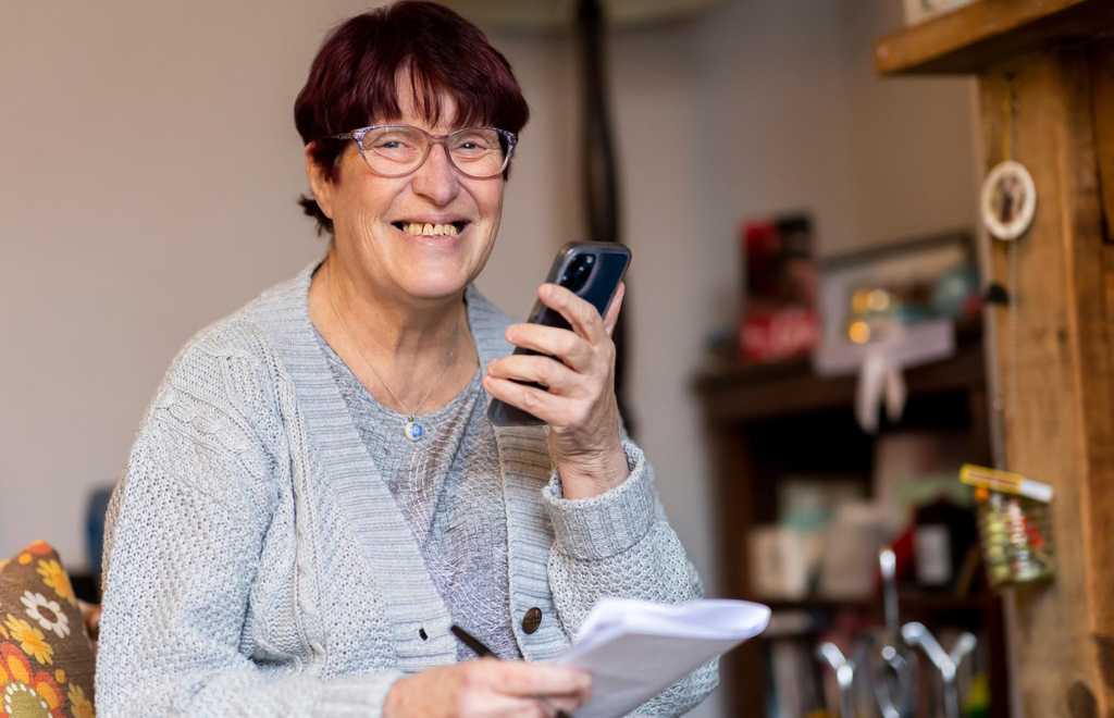 A woman stands in a kitchen smiling and as she talks on her phone which she is holding in front of her.