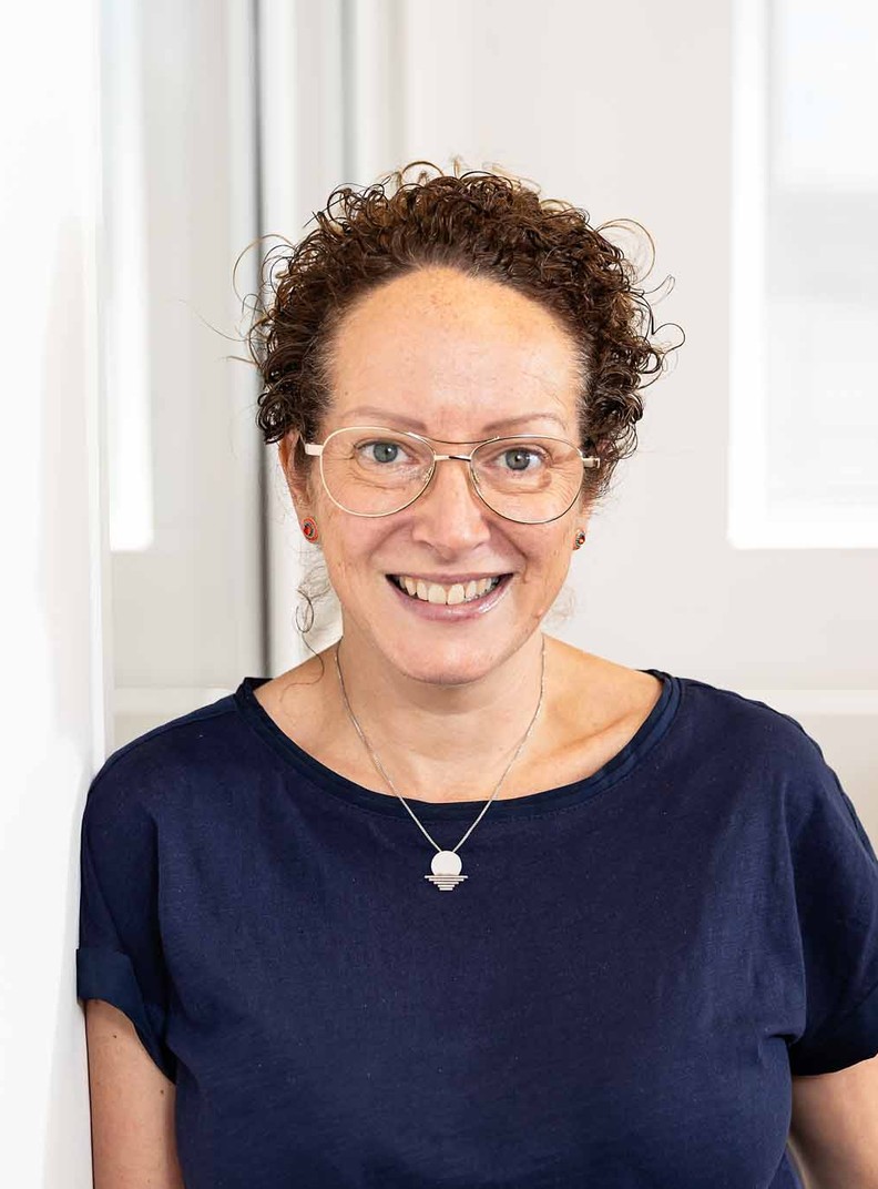 Julie Hocking, Individual Giving & Legacies Manager for BWBF, leans against a wall and is smiling. She wears glasses and her curly hair is tied up.